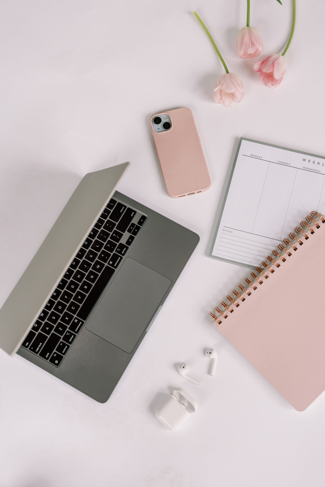 A desk with a laptop, half open, iphone with pink case, pink notebook, all scattered throughout the white desk.