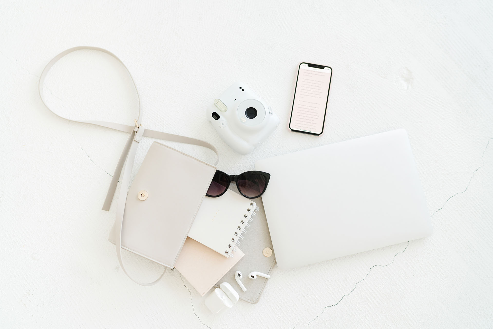 A flat lay of minimalist accessories including a white camera, smartphone, laptop, sunglasses, and a beige purse, arranged on a white surface.